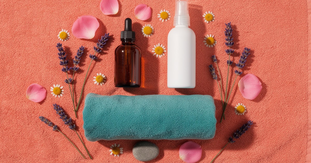 An overhead flat lay on coral with an amber dropper bottle, a white spray bottle, a rolled teal towel, lavender stems, daisies, pink petals and a gray stone.